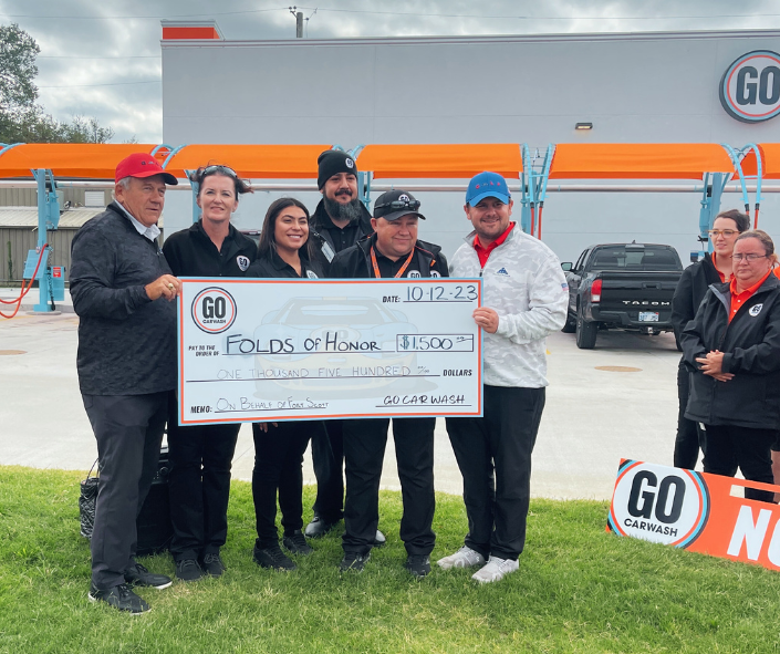 Group photo at a GO Carwash location featuring staff and a representative holding a large ceremonial check for $1,500 made out to "Folds of Honor." The check is dated 10-12-23, and the donation is noted to be on behalf of "Folds Sweep." The background shows car wash equipment and branding.