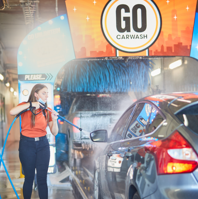 A GO Carwash employee using a high-pressure water hose to pre-wash a car before it enters the automated wash system. The car is positioned at the entrance of the car wash tunnel, with the GO Carwash sign and blue foam brushes visible in the background