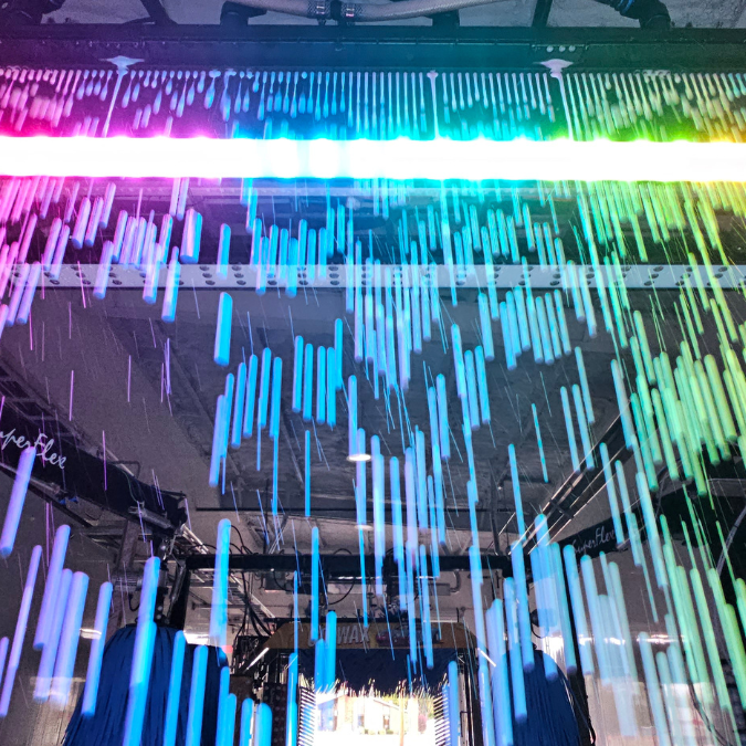 Vibrant image from inside a car wash tunnel, featuring colorful LED lights creating a rainbow effect as water sprays down. The lights and water droplets form dynamic patterns, adding an exciting visual experience to the car wash process.